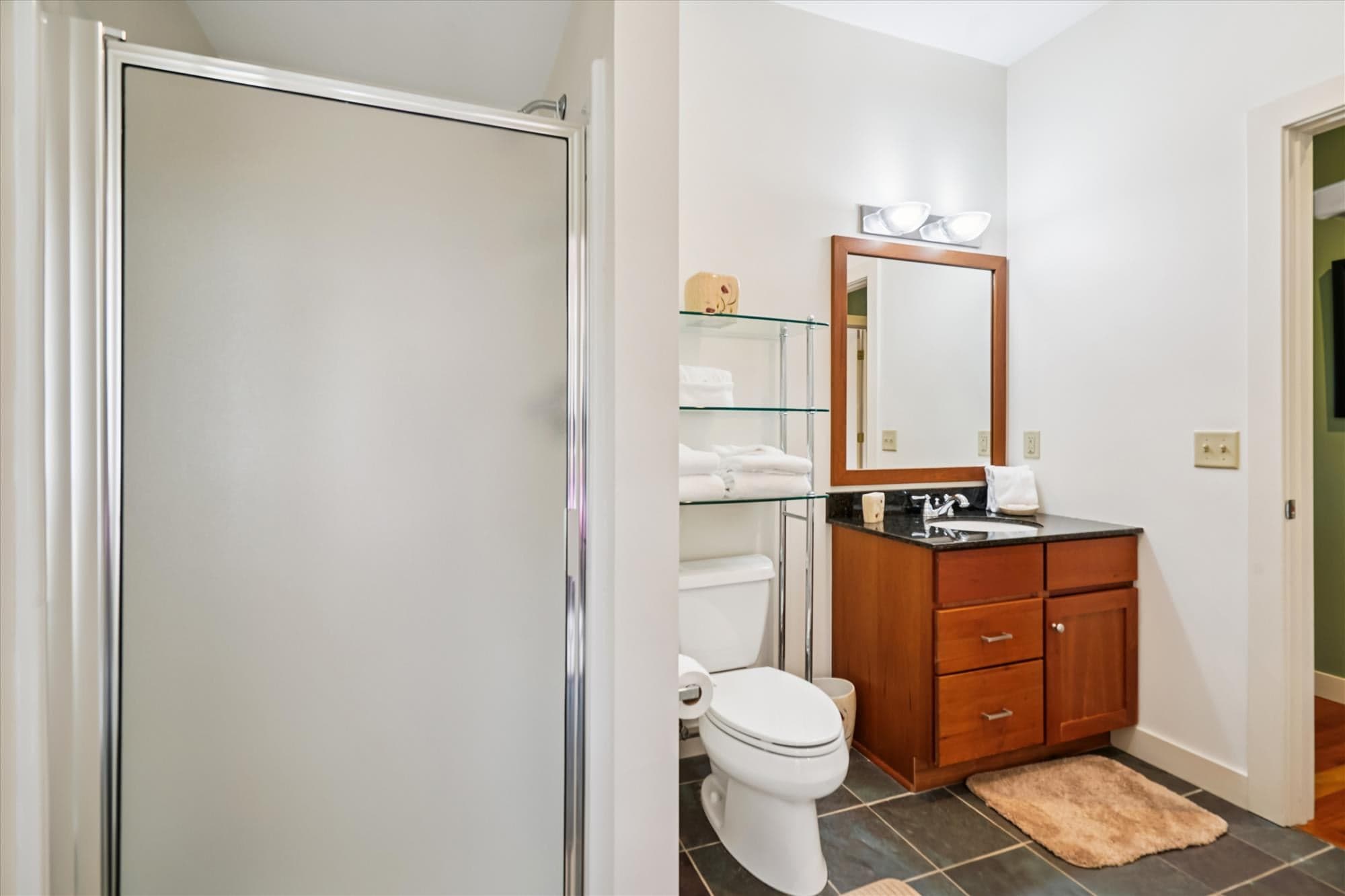 A bright full bath featuring a walk-in shower, granite vanity, and glass shelving stocked with fresh towels. Conveniently located near the queen bedroom for privacy and comfort.