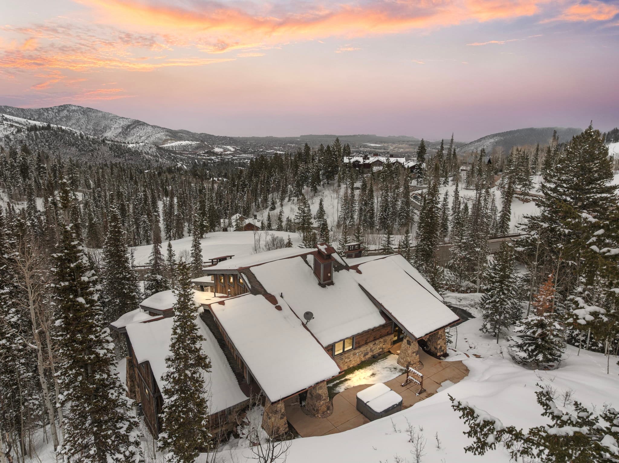 rear view of the homes exterior, there is a bright sunset and snow covered mountains in the background, you can see the hot tub and outdoor living area